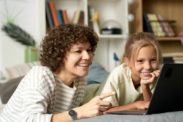 Happy mother watching funny video on laptop together with her daughter, they looking at monitor and laughing