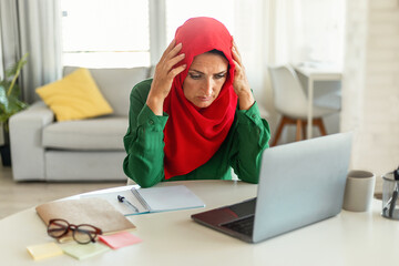 Wall Mural - Concerned mature islamic woman using laptop, reading negative news online and touching head, sitting at home