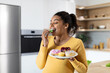 Smiling hungry young black pregnant female with big belly eats sweets, hold plate with cookies