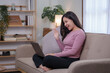 Beautiful Asian woman sitting on sofa at home with laptop and headphones on video call chatting and smiling brightly for today.