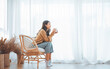© oatawa - Happy asian woman relaxing drinking hot coffee or tea in holiday morning vacation on armchair at home, Cosy scene, Smiling pretty woman drinking hot tea in autumn winter. copy space.