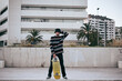 caucasian young man in striped sweater and nice hat standing looking at camera with his skateboard in hand on the city street