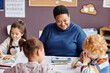 © Seventyfour - Focus on happy African American mature teacher and little learner with dark long hair sitting in front of two intercultural nursery schoolkids