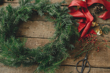 Wall Mural - Making Christmas wreath. Fir branches on round christmas wreath, red ribbon, berries, scissors on rustic wooden table. Moody holiday image. Holiday workshop, winter decor