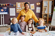 © Seventyfour - Happy mature teacher and group of intercultural little learners standing by desk and looking at camera at lesson in classroom