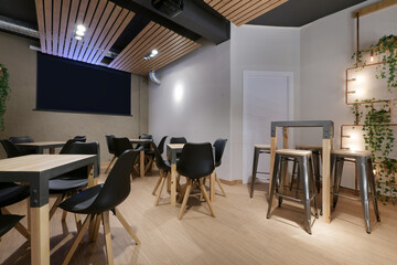 Hall of a restaurant with wooden tables and black resin chairs and a movie screen in the background