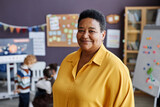 Successful professional mature female teacher of nursery school looking at camera while standing against group of little learners