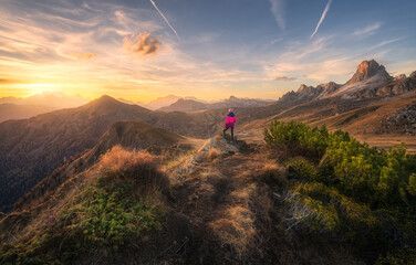 Wall Mural - Young woman on the stone on mountain peak and beautiful mountains at colorful sunset in autumn. Dolomites, Italy. Sporty girl, mountain ridges, orange grass, green trees, golden sun in fall. Hiking	