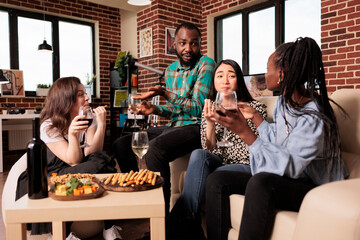 Culturally diverse friends talking, conversing each other, drinking wine, eating, tasting cheese, bread sticks at diverse people gathering. Different nationalities mates enjoying social life.