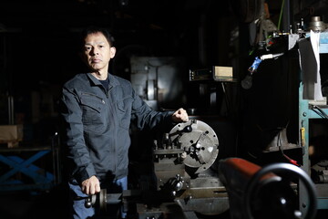 A craftsman poses in gray work clothes in front of a lathe at a local factory. Conceptual images of the essence of manufacturing, technical succession, and the challenge of high-precision machining.