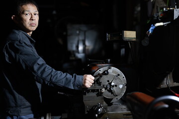 A craftsman poses in gray work clothes in front of a lathe at a local factory. Conceptual images of the essence of manufacturing, technical succession, and the challenge of high-precision machining.