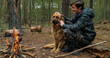 Young man with big dog sits at the fire in the forest. Cheerful guy is stroking his dog, outdoor. Happy caucasian teenager with his German shepherd by the fire in nature, in autumn.