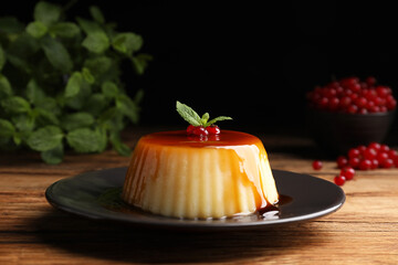 Plate of delicious caramel pudding with red currants and mint on wooden table against black background