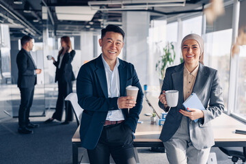 Wall Mural - Two happy multiracial colleagues talking and drinking coffee during break time in office