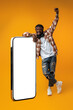 Smiling african man standing near huge smartphone with blank screen on yellow studio background