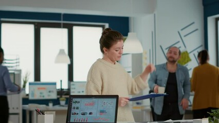 Wall Mural - Casual woman asking coworker for help finding paper with charts in front of laptop with business software standing at desk. Startup employee talking with colleague about sales in busy office.