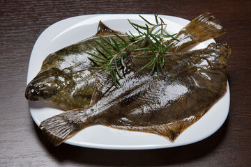 Two raw flounders and sprigs of rosemary on a plate, on a brown table