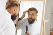 © Studio Romantic - Morning hygiene. Handsome stylish man combing and styling his hair in morning in front of mirror. Bearded millennial man in bathrobe looks at his reflection in mirror and takes care of himself.