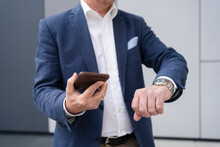 Businessman Holding Mobile Phone And Checking Time On Wrist Watch In Front Of Gray Wall