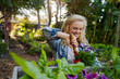 © StratfordProductions - Young caucasian woman in checked shirt using spade on flowers near plants in plant nursery