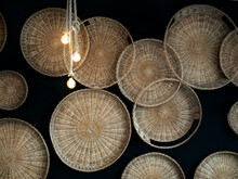 Background Composed Of Wicker Trays Hung On A Black Wall. Light Bulbs Suspended By Ropes Partially Illuminate The Scene.