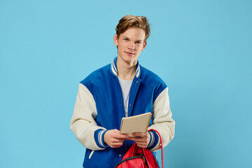 Wall Mural - vertical portrait of a handsome pleasant red-haired guy in a fashionable blue jacket with a red backpack and an empty notebook in his hands on a blue background in the studio