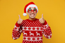 Merry Satisfied Fun Young Man Wear Knitted Christmas Sweater Santa Hat Glasses Posing Show Thumb Up Like Gesture Isolated On Plain Yellow Background. Happy New Year 2023 Celebration Holiday Concept.
