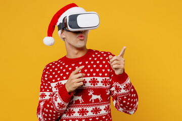 Wall Mural - Merry young man wear red knitted christmas sweater Santa hat posing watching in vr headset pc gadget point finger aside on area isolated on plain yellow background Happy New Year 2023 holiday concept.