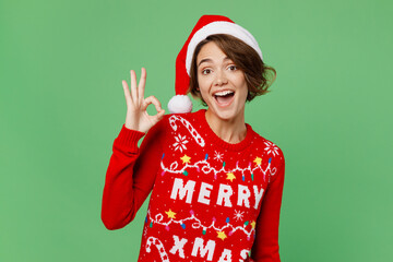 Merry smiling fun young woman 20s wear knitted xmas sweater Santa hat posing showing okay ok gesture isolated on plain pastel light green background. Happy New Year 2023 celebration holiday concept.