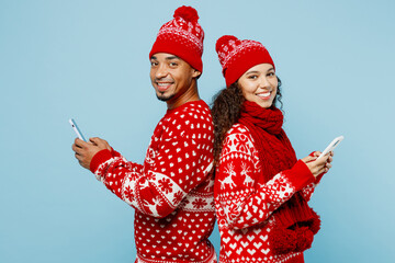 Sideways fun merry young couple two man woman wear red Christmas sweater Santa hat posing hold use mobile cell phone isolated on plain pastel light blue background Happy New Year 2023 holiday concept