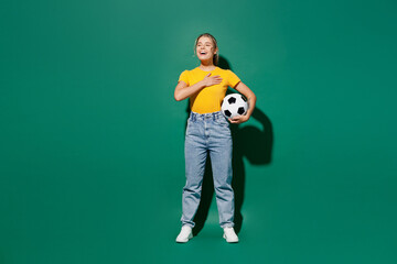 Full body young woman fan in basic yellow t-shirt cheer up support football sport team hold soccer ball watch tv live stream put hand in heart sing hymn song isolated on dark green background studio