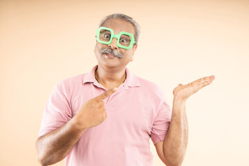 Wall Mural - Excited Senior indian man wearing funky sun glasses pointing finger for advertisement isolated on beige background, blank space for promotion, look here. Funny old male.