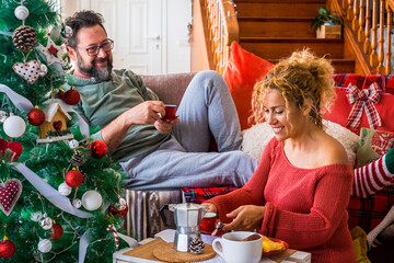 Wall Mural - Happy couple family man and woman enjoy christmas holiday morning at home having breakfast together. Xmas tree and coffee time. December event season. Mature lady and husband enjoy indoor leisure