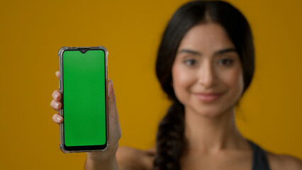 Indian ethnic woman lady girl lady in yellow studio background showing smartphone mobile telephone screen with green empty chroma key touchscreen technology cell message display advertise web service