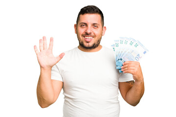 Young caucasian man holding a banknotes isolated smiling cheerful showing number five with fingers.