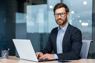 Wall Mural - Portrait of mature successful and happy businessman, senior man with beard smiling and looking at camera, investor working with laptop inside office in business suit.