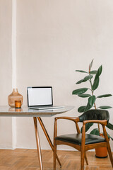 A minimal interior scene with blank laptop screen mockup, candle, vase on the table, potted plant and stylish chair. Modern inspiring workplace.