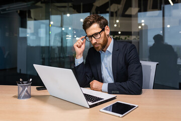 Wall Mural - Serious thinking senior businessman working inside office using laptop, man in glasses solving problem, mature investor in business suit.