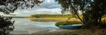 Serene Morning Landscape. Panoramic View From The Shore Of A Large Lake With A Fishing Boat, Sky Reflection On The Water, A Forest On The Opposite Shore And A Frame Of Tree Branches