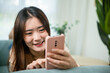 © sorapop - Smiling attractive woman relaxed reading text message on smartphone, Asian young female using smart mobile phone lying on sofa in living room room at home, gadget online application making internet