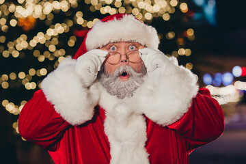 Wall Mural - Portrait of impressed santa claus pouted lips arms touch glasses staring cant believe midnight tree garland city center outside