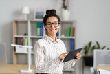 Portrait of happy female office worker holding digital tablet, smiling at camera, standing near her workplace in office
