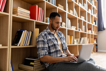 Poster - Young arab guy studying online from home, using laptop pc, sitting on floor near bookshelves, copy space