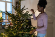 Young happy smiling african woman decorating Christmas tree in minimalist way, cheerful black girl stringing LED garland along branches, preparing your home for holiday season. shallow depth of field