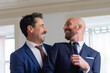 © KAMPUS - Homosexual couple in official suits smiling each other. Happy Caucasian gay couple getting dressed for wedding ceremony in hotel room, enjoying each others company. LGBT, love, marriage concept