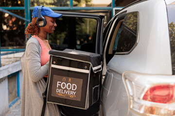 African american smiling courier delivering takeaway food by car, waiting for customer near office building. Delivery service worker taking out backpack from vehicle, side view