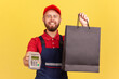 © khosrork - Portrait of bearded happy positive courier man bringing order in black shopping bag and giving pos terminal to client to pay for purchase. Indoor studio shot isolated on yellow background.