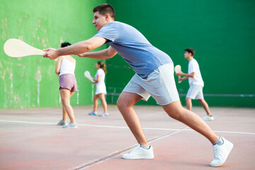 Portrait of emotional determined young guy playing pelota at open-air fronton in summer, swinging traditional wooden bat to return ball. Sportsman ready to hit volley