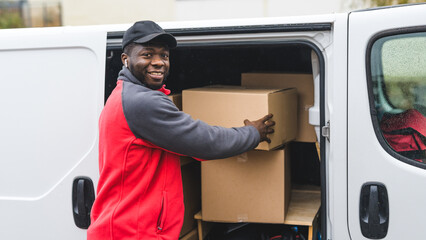 Wall Mural - Upcoming holiday season sale via online shopping. Outdoor medium closeup shot of smiling surprised Black man in red-and-gray working clothes taking out one of many cardboard boxes out of his white van