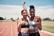© L Ismail/peopleimages.com - Portrait, success medals and women at stadium after winning running race or sports event outdoors. Fitness, winner and athlete friends happy with victory, goals or target achievement on track field.